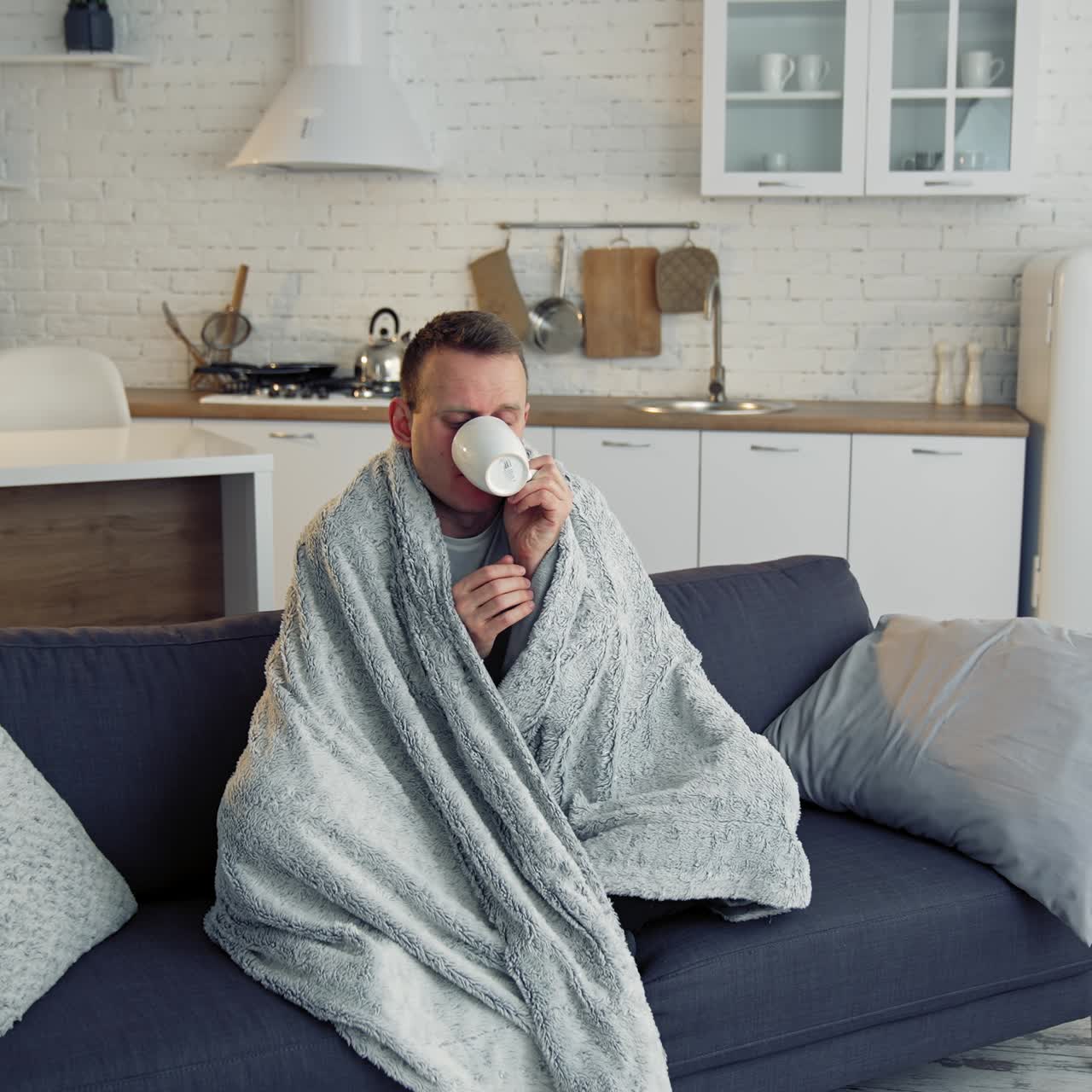 Ill man drinking hot beverage at home. Unwell young man covered with warm blanket sitting on a sofa in the kitchen. Coronavirus pandemic