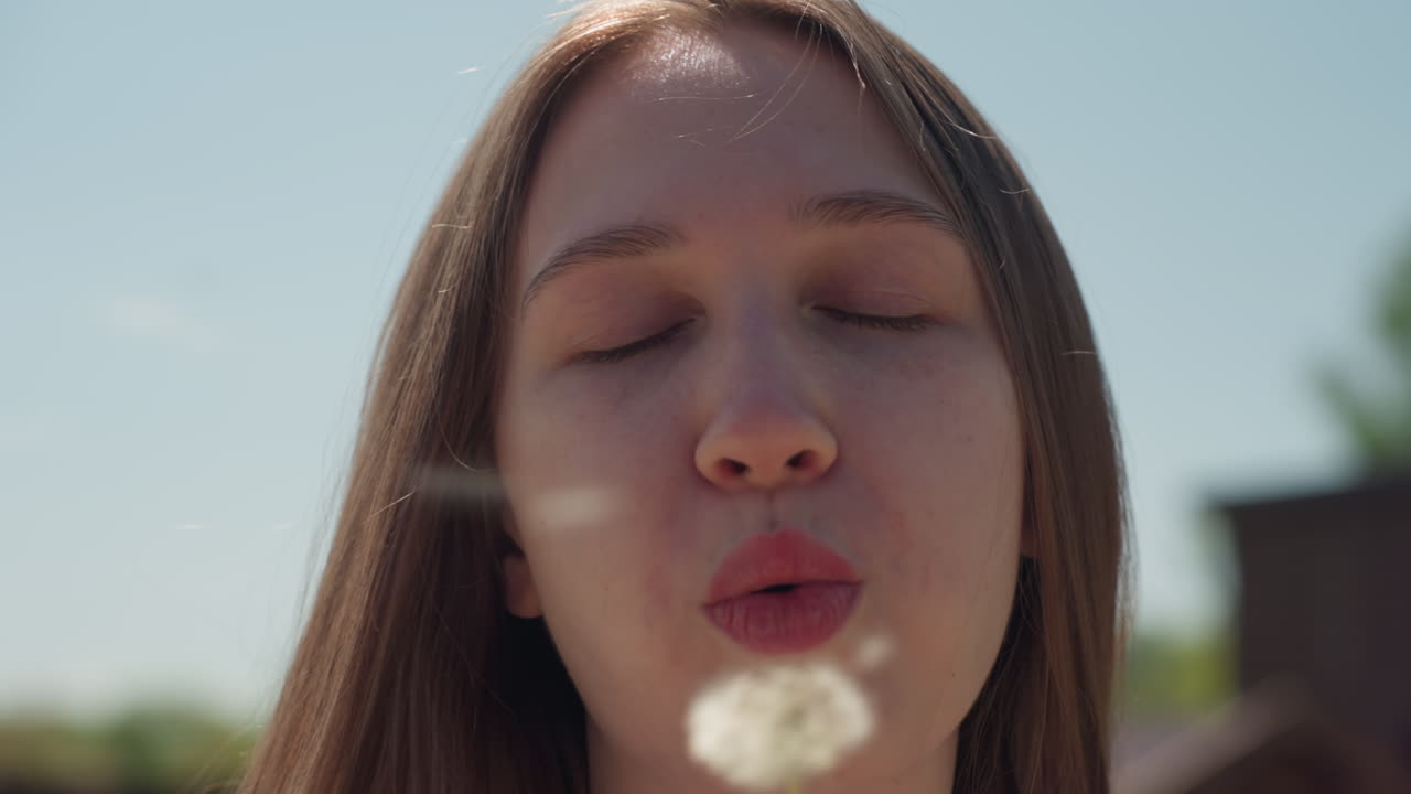 Young woman with long hair gently blows dandelion under bright sunlight, holding stem close to lips with calm expression, surrounded by clear blue sky, distant rooftops, and natural outdoor setting