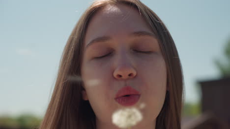 young woman with long hair gently blows dandelion under bright sunlight, holding stem close to lips with calm expression, surrounded by clear blue sky, distant rooftops, and natural outdoor setting