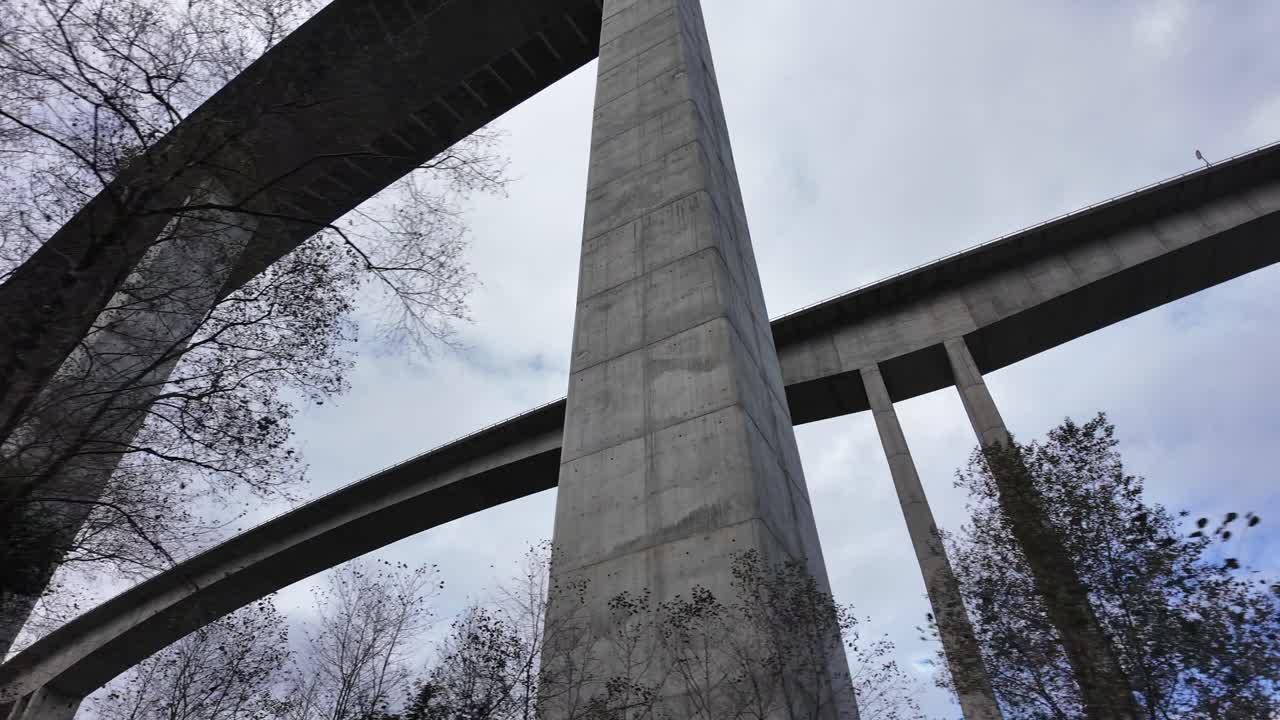 A massive highway viaduct rises above a forested gorge from a dramatic low-angle view. Filmed at the Viaducto de La Concha de Artedo on the Autovía del Cantábrico, Asturias