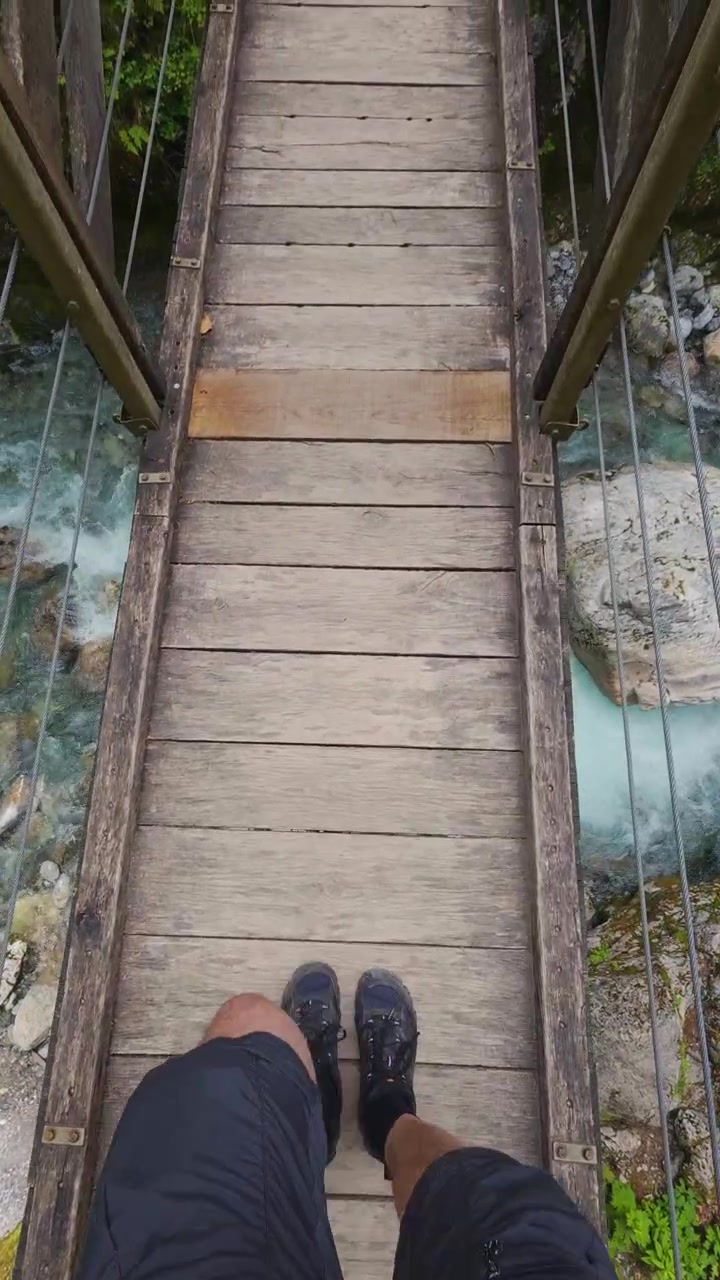 POV verticale, attraversando un ponte sospeso di legno sopra il fiume Isonzo turchese in Slovenia, mostrando calzature da trekking, assi del ponte, acqua con rocce e paesaggio di viaggio all'aperto.