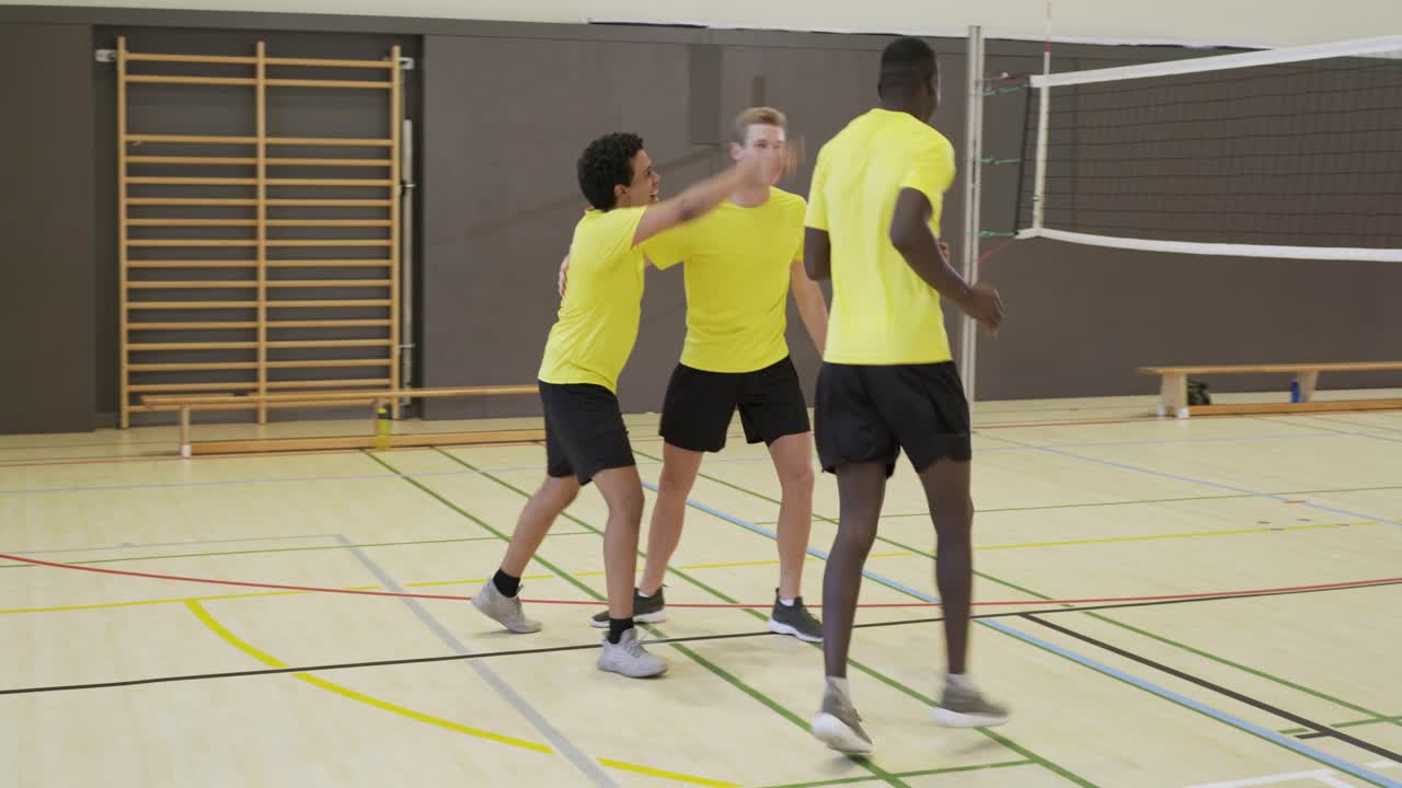 Tres jugadores masculinos con camisetas amarillas celebrando después de que el oponente no lograra devolver un remate de balón en el gimnasio.