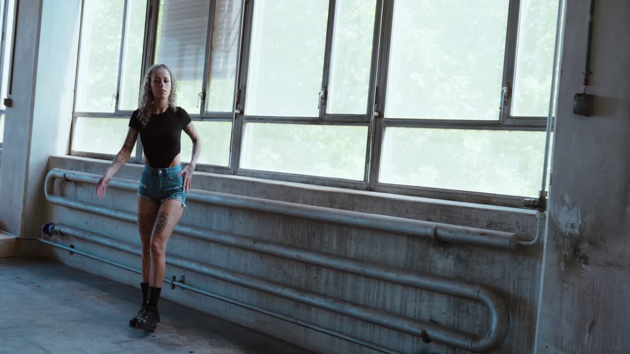 Tattooed woman posing in a warehouse next to a window, she wears hot pants, slow motion