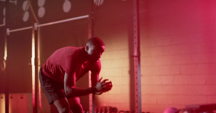 Uomo afroamericano che si allunga verso la scarpa, accovacciato, mentre si riscalda vicino a kettlebell sotto l'illuminazione rossa della palestra.