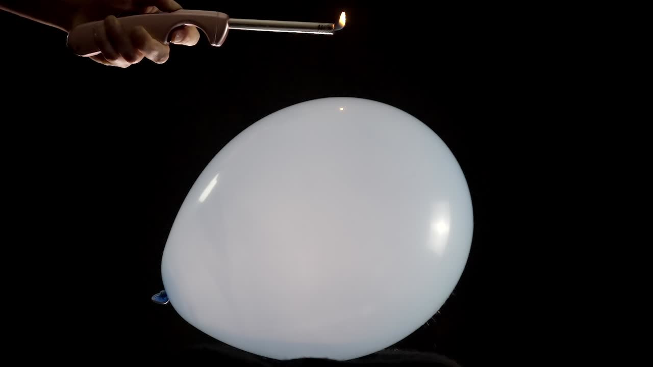 A big balloon is displayed as a hand holding a lighter moves closer. The tension builds in slow motion, culminating in the dramatic moment when the balloon pops, creating an intense visual effect.