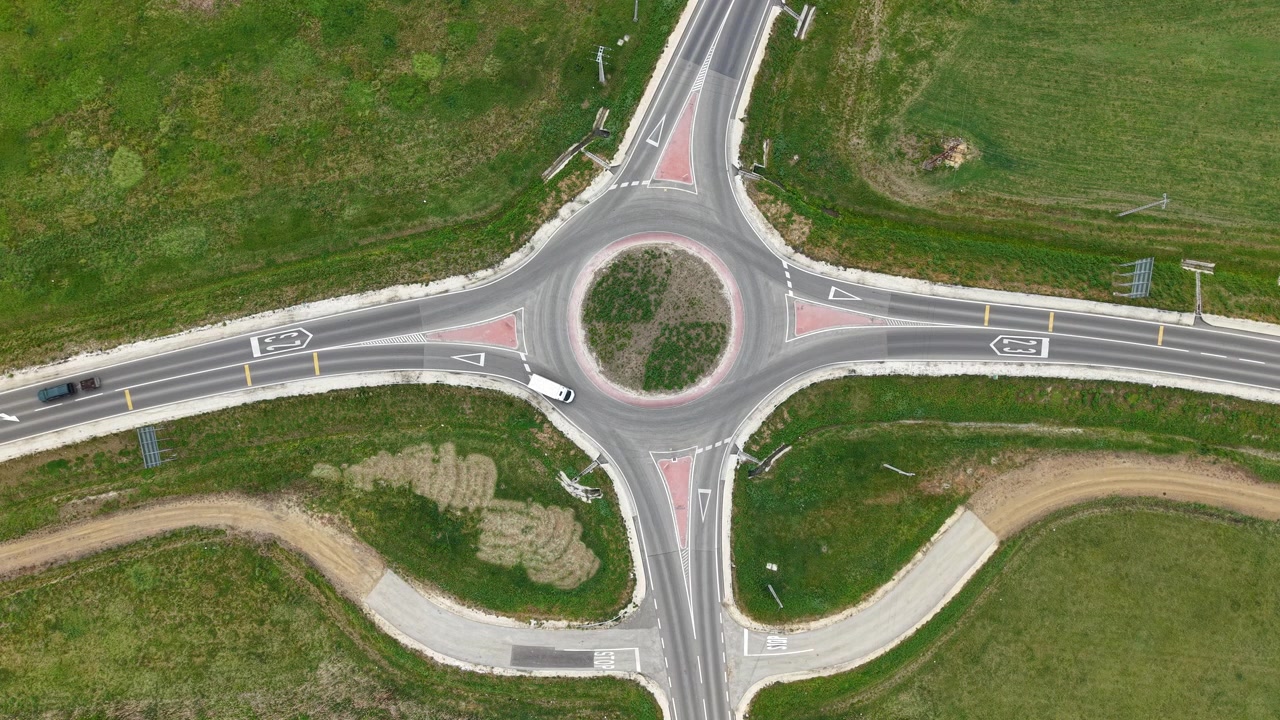 An aerial view captures vehicles traveling through a paved roundabout intersection located in a scenic rural green landscape in daylight.