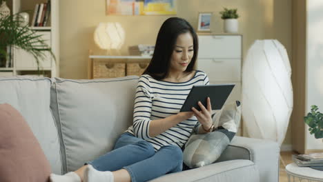 Young-Woman-Watching-Something-On-The-Tablet-And-Tapping-On-The-Screen-Sitting-On-A-Sofa-In-The-Living-Room-1