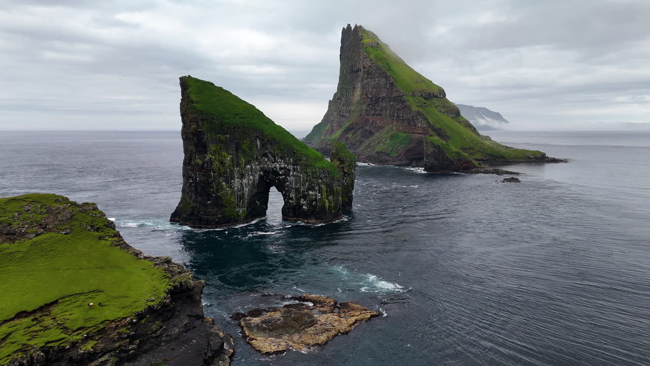 Cinematic aerial view of Drangarnir sea stacks rising dramatically from the Atlantic Ocean near Vágar, Faroe Islands, showcasing rugged cliffs, lush green slopes, and misty Nordic seascape