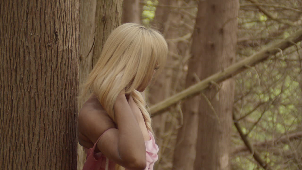 Young ebony woman, wearing a pink satin gown, leaning against trunk of a Sweetgum tree.. She plays with her blonde hair, before looking at camera, then up at the sky. Medium Shot. 60fps slow motion.