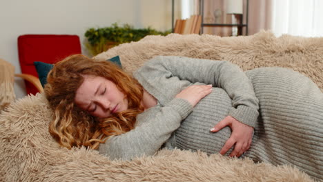 pregnant young woman lying on home sofa holding belly wincing in pain discomfort worried expression