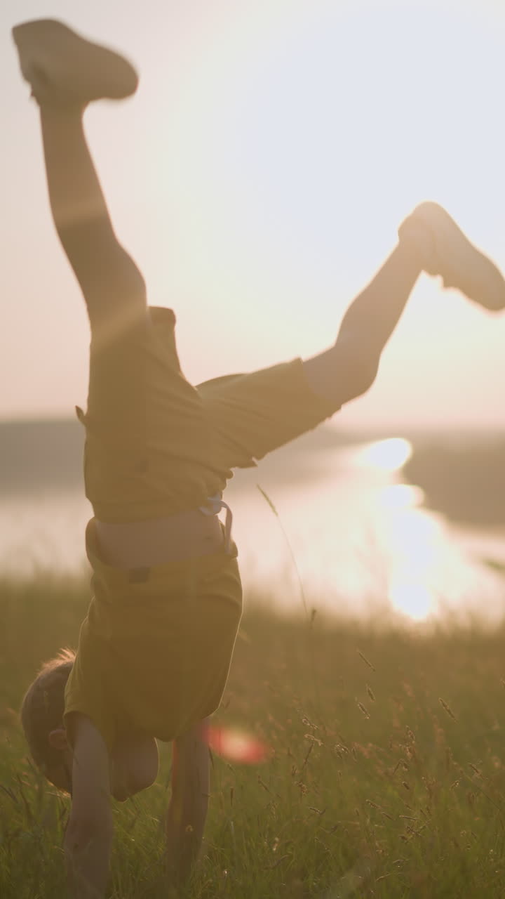 A young boy, dressed in yellow and wearing glasses, is caught mid-flip in a grassy field during a stunning sunset. The scene captures his playful energy, with a serene lake visible in the background