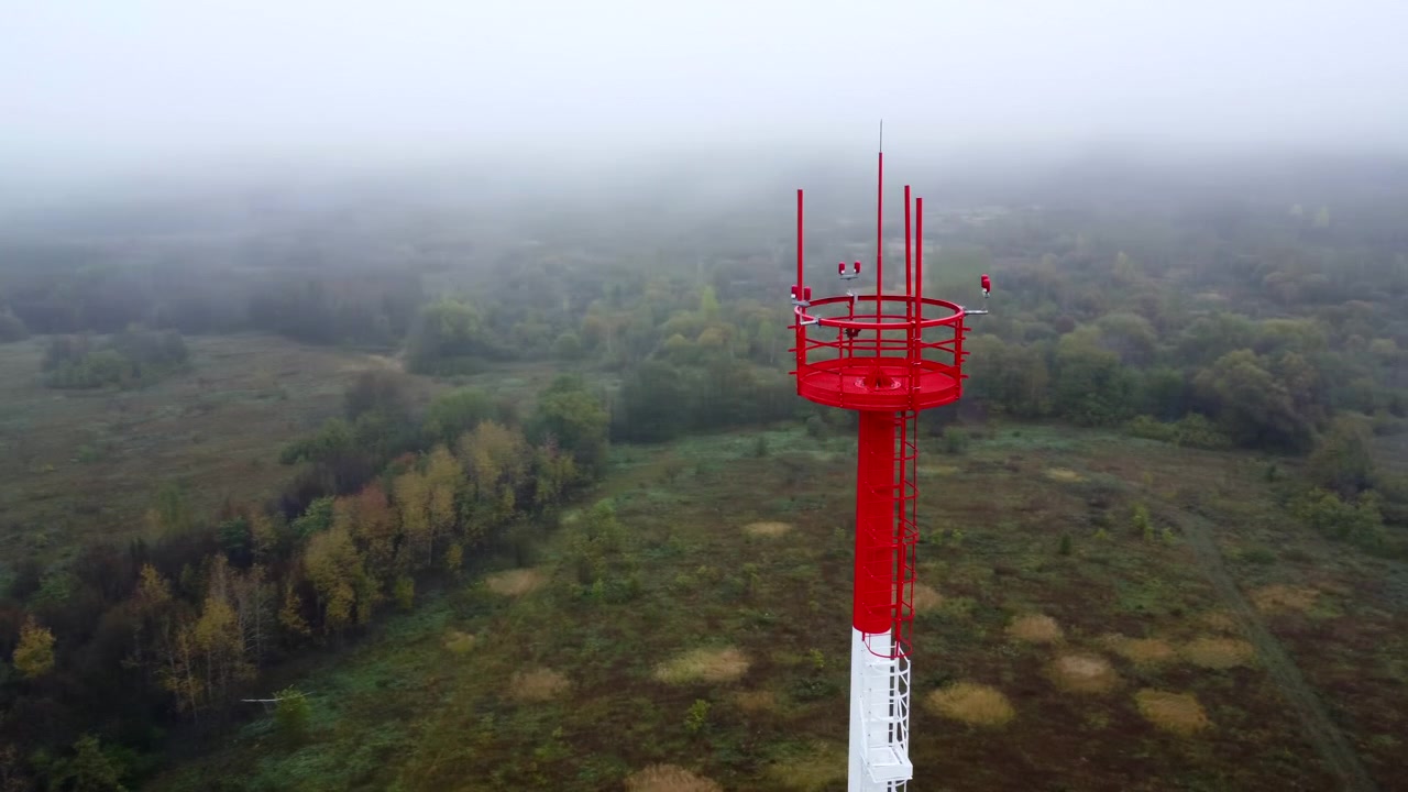 Un'alta torre di comunicazione rossa e bianca si erge sulla destra sopra un paesaggio rurale nebbioso, con alberi fitti, campi aperti e una luce soffusa e nuvolosa che svanisce