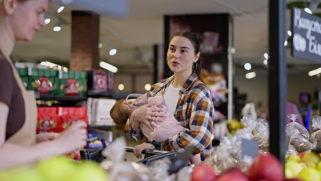 una feliz chica morena con un bebé en los brazos le pregunta a un trabajador del supermercado sobre la calidad de los productos y qué es mejor elegir durante sus compras