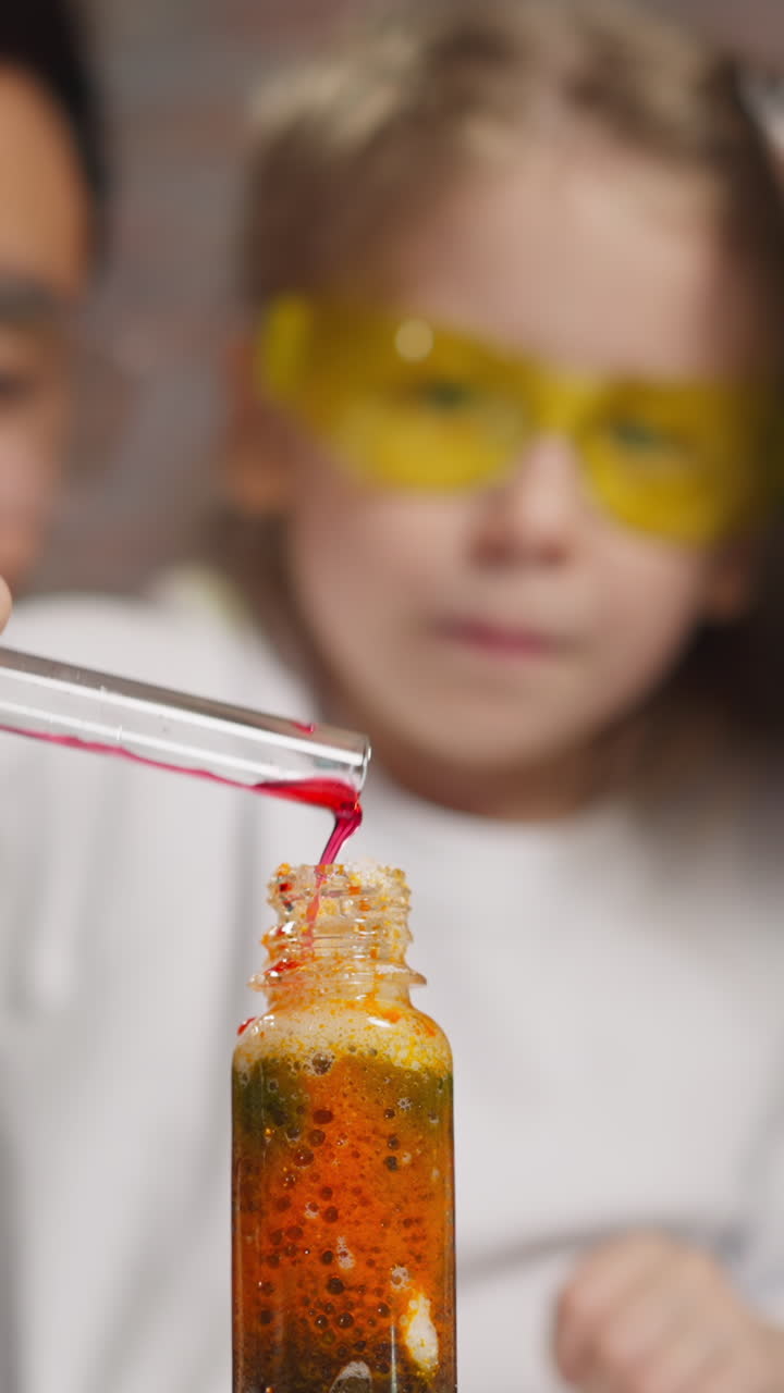 Playful little girl licks lips and pours red liquid into bottle with foamy substance conducting experiment between teachers at lesson closeup