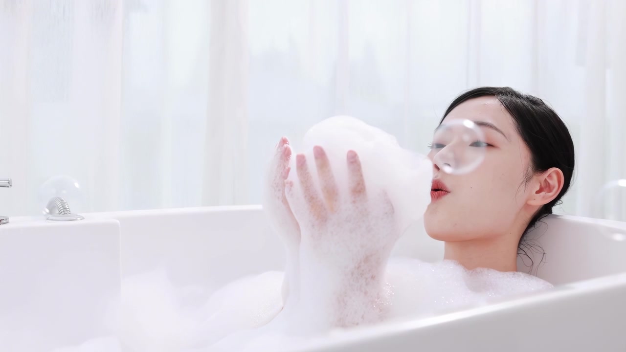 Bella giovane donna rilassata, sorridente e seducente, avvolta in un asciugamano, che soffia schiuma mentre si lava seduta nella vasca da bagno. La ragazza si gode un bagno di lusso con bolle e sapone per una cura perfetta della pelle