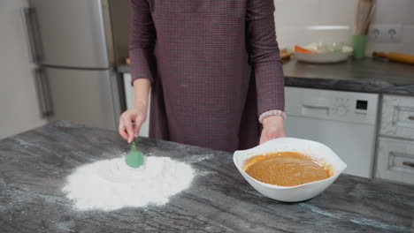 baker uses spatula to create hole in sifted flour on countertop, preparing to pour creamy batter, kitchen setting with utensils and ingredients