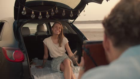 pov: a bearded man with curly hair in a blue shirt is taking photos of his blonde girlfriend, who is smiling and playing the ukulele in the trunk of the black car decorated with the lights against the yellow sky