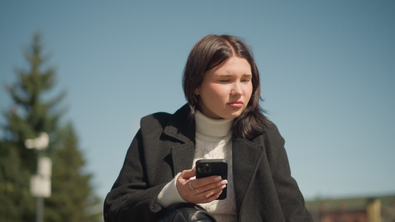 молодая женщина, сидящая на скамейке на открытом воздухе, смотрит в даль, держа в руках смартфон, фокусируется на своем телефоне назад, в черном пальто и белом свитере, солнечный свет освещает ее лицо