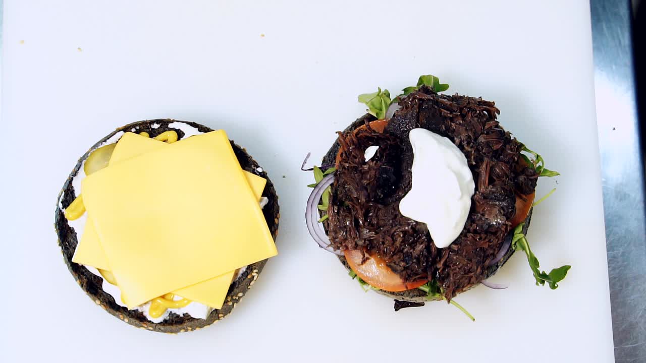 Cooking healthy hamburger. Chef puts white sauce with a spoon on burger with vegetables. Halves of black buns with tasty ingredients for making vegan burger. Top view