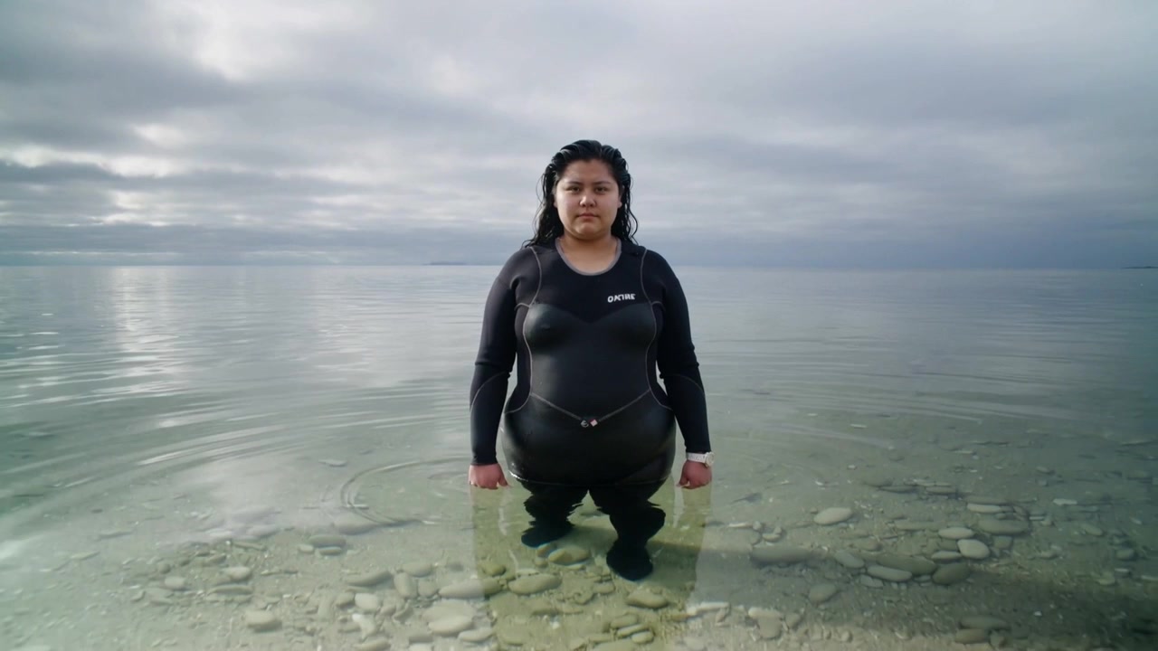 Standing Hispanic woman in black wetsuit shifting feet at pebble shore, camera pulling back