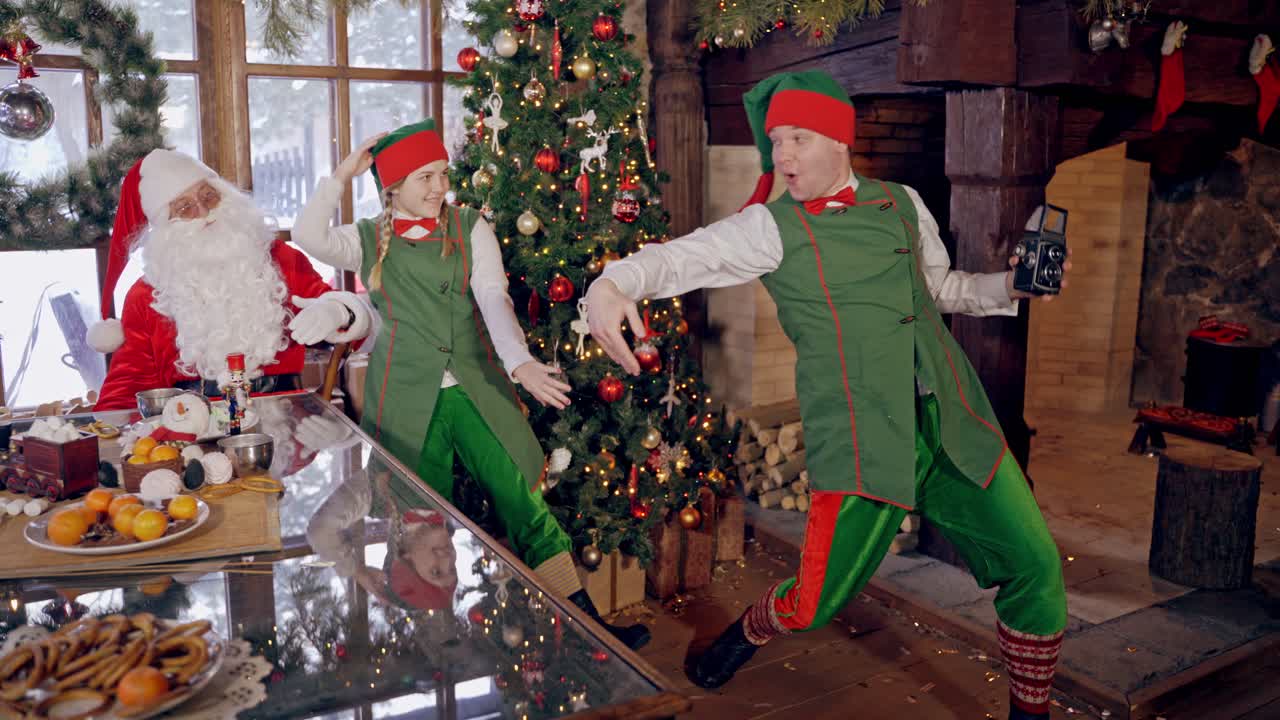 Santa together with elf near the Christmas tree. Funny elf is taking photo of fairy characters in Christmas decorated room.