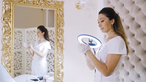 an attractive permanent makeup master prepares for the procedure, wearing white latex gloves near a huge mirror with golden trim in the beauty salon.