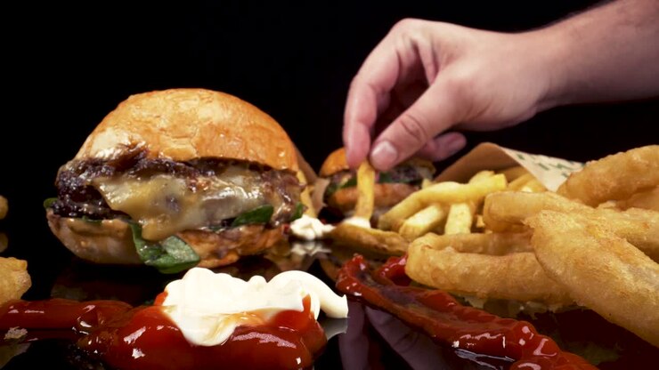 Moving Macro view passing through fast food, burgers, fries, onion rings, ketchup and mayo on black reflective glass, hands picking up food to eat
