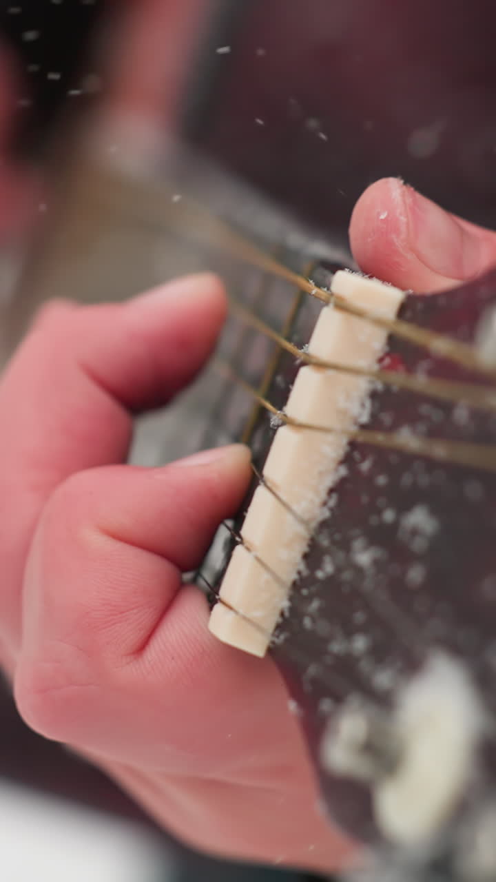 겨울의 설정에서 기타 스트링을 연주하는 손의 클로즈업, 스트링과 손가락판의 얼어붙은 세부 사항을 강조, 손의 따뜻함과 차가운 눈 바탕 사이의 생생한 대조