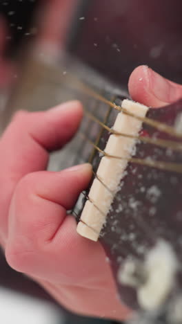 primer plano de la mano tocando cuerdas de guitarra en un entorno de invierno, destacando los detalles helados en las cuerdas y la pizarra, un vibrante contraste entre el calor de la mano y el frío fondo nevado