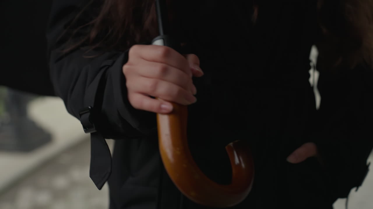 Nærbillede af træparaplyhåndtag, skræddersyet sort frakke, dekorativt altangelænder, diskret bevægelse, der antyder gang eller venten, urbant morgenlys, cinematisk farve, blød bokeh, fortælling