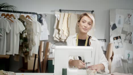 mujer sastre parada en el taller de costura, luego se sienta a coser con una máquina de coser y mira la cámara