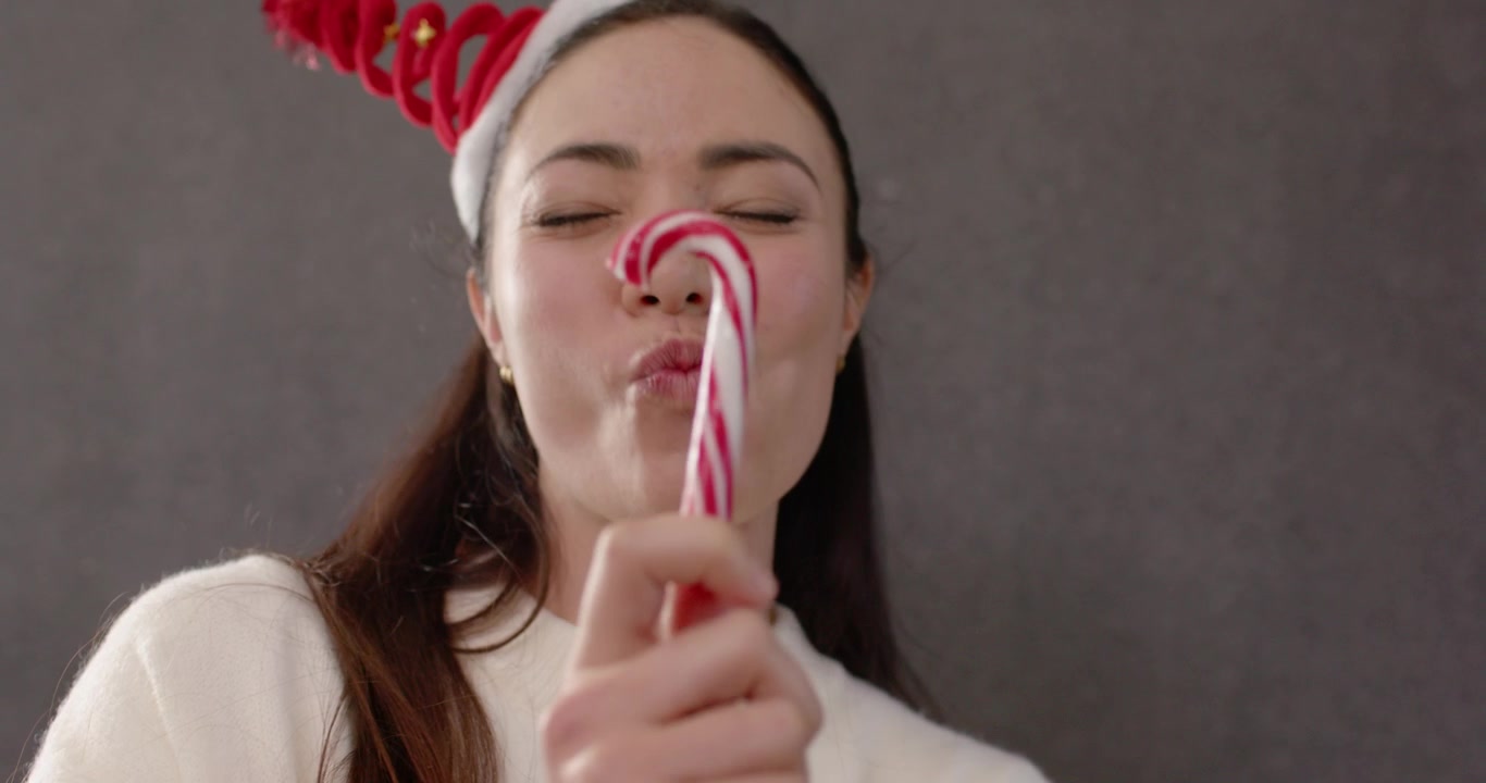 Donna che soffia in una trombettina da festa, tira e bacia una caramella a bastoncino, festeggiando in una stanza con maglione bianco e fascia rossa.