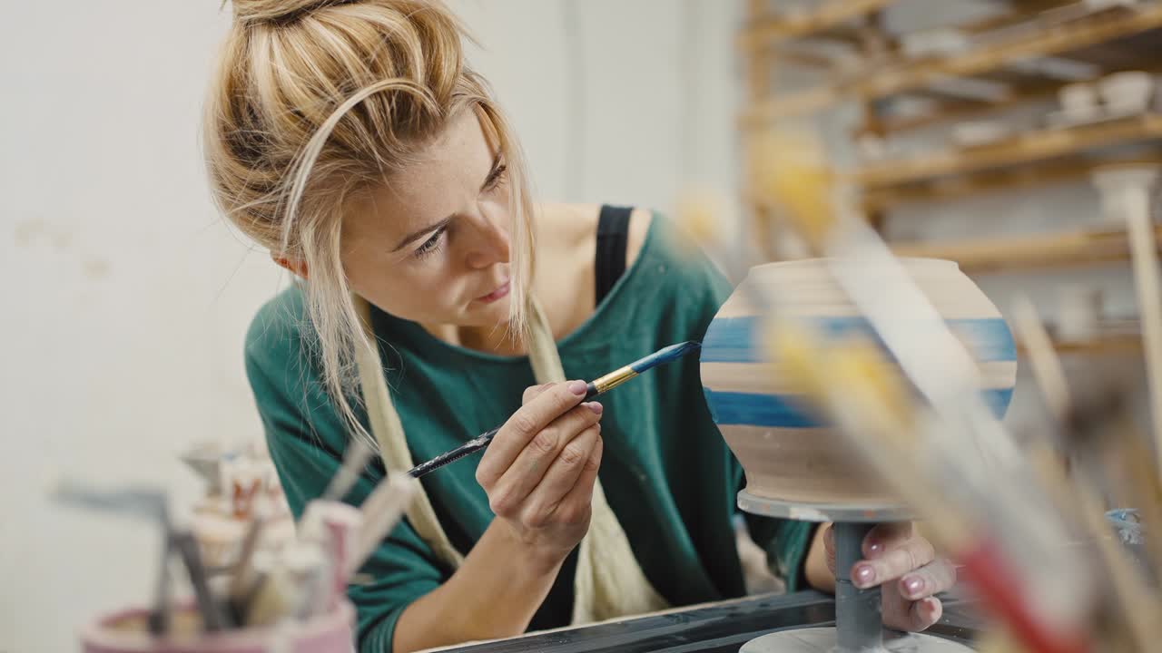 Focused woman artist painting handmade ceramic pot with blue dye, sitting at pottery workshop, slow motion