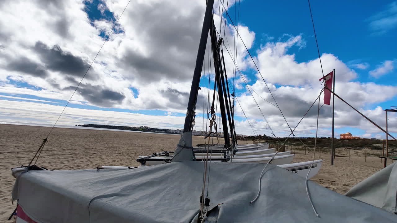 A detailed view of sailboat ropes and masts on a quiet beach, with a backdrop of a cloudy sky and a distant coastline. Small boats on the beach. In Algarve, Portugal.
