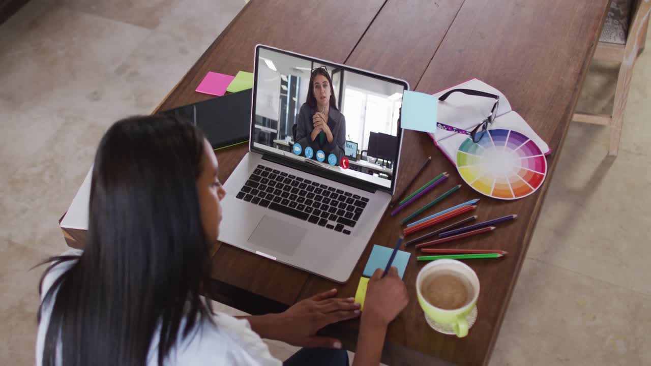 Mujer afroamericana escribiendo en notas adhesivas mientras hace una videollamada en su portátil en casa.
