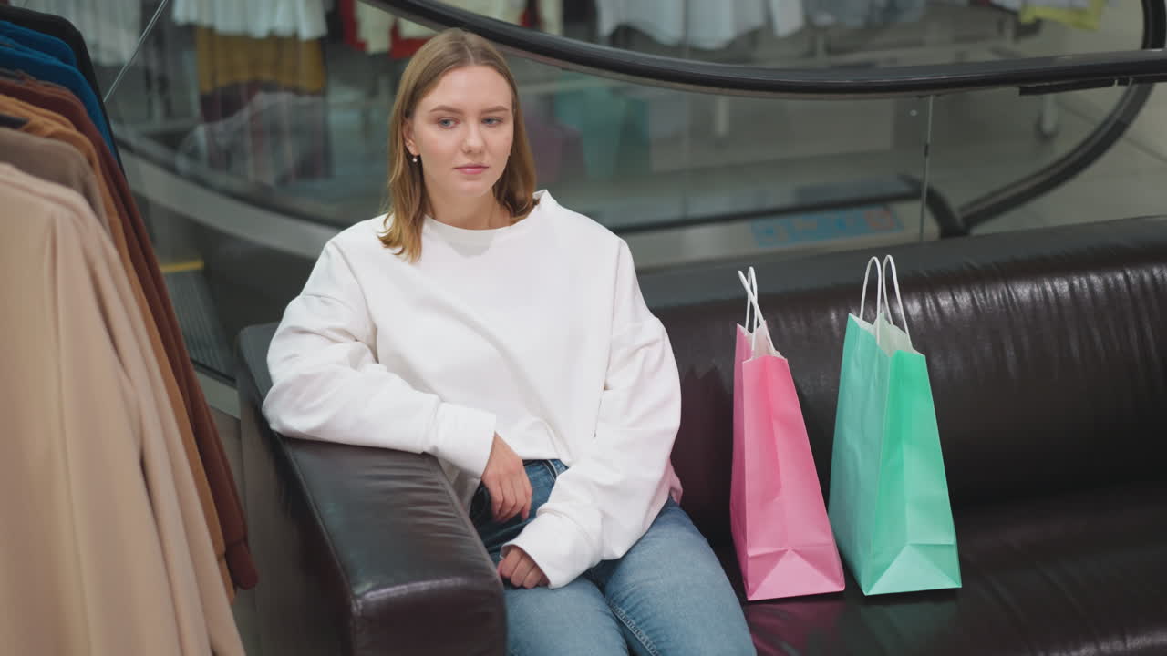 현대 쇼핑몰에서 가죽 의자에 앉아있는 자신감있는 여성, 캐주얼 하 과 청바지를 입고, 유리 에스컬레이터 배경으로 다채로운 쇼핑 가방과 함께