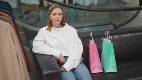 dama confiada sentada en una silla de cuero en un centro comercial moderno, vestida con una camiseta blanca casual y vaqueros, acompañada de bolsas de compras de colores, con fondo de escalera mecánica de vidrio