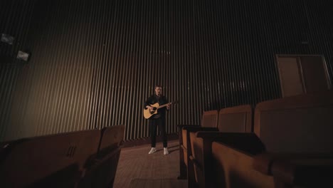 a handsome guy plays an acoustic guitar in an abandoned cinema. the musician sings a song and accompanies on the guitar