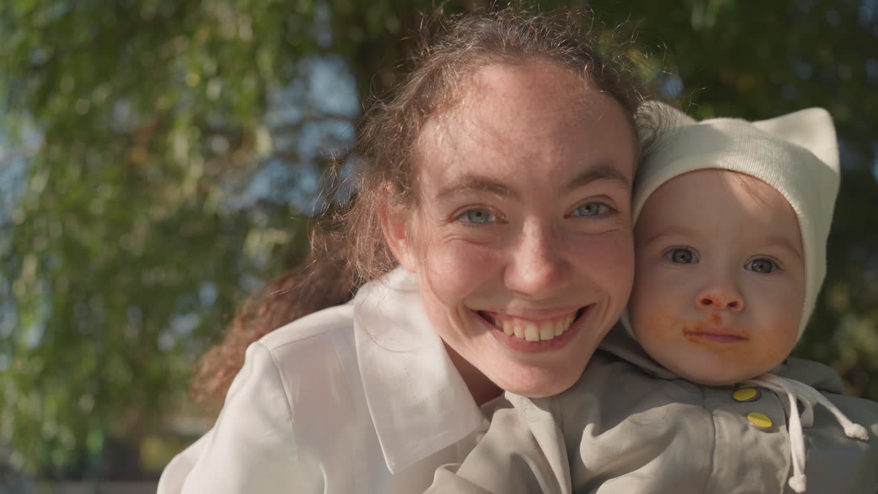 Glædeligt familiemoment. Nærbillede af smilende mor og baby. Solbeskinnet park indfanger glad familie, der nyder samvær. Levende billede af smilende mor og baby i et lyst, solrigt parkmiljø.