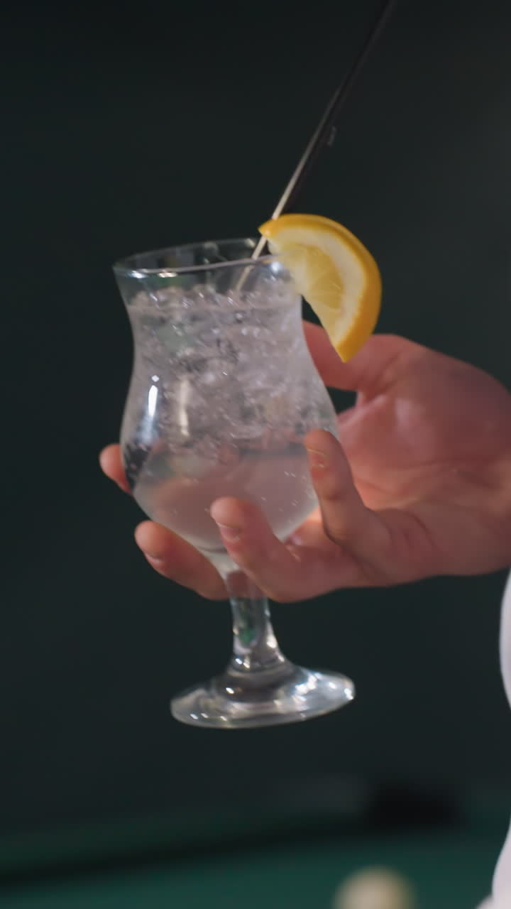 Primo piano di una mano che tiene una stecca da biliardo e una limonata mentre cammina in una sala da biliardo. I cubetti di ghiaccio e la fetta di limone nel bicchiere creano un contrasto rinfrescante sullo sfondo scarsamente illuminato con il tavolo da biliardo verde.