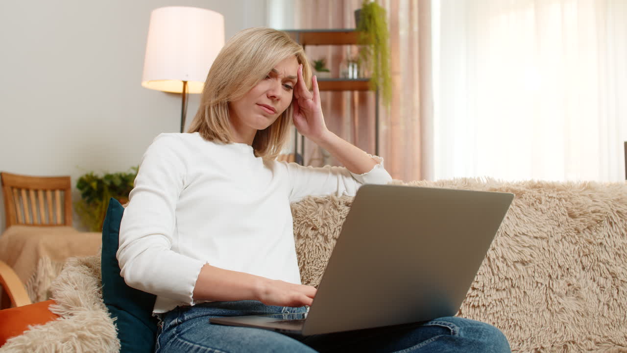 Woman massaging temples on sofa at home feeling tired from long work on laptop headache discomfort
