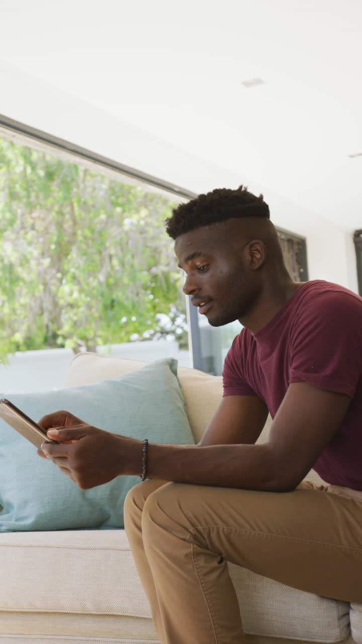 Vertical video of happy african american man using tablet at home