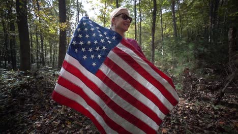 Hübsche-Blonde-Frau,-Die-Sich-In-Eine-Flagge-Hüllt,-Während-Die-Kamera-Sie-In-Einem-Wald-Umkreist