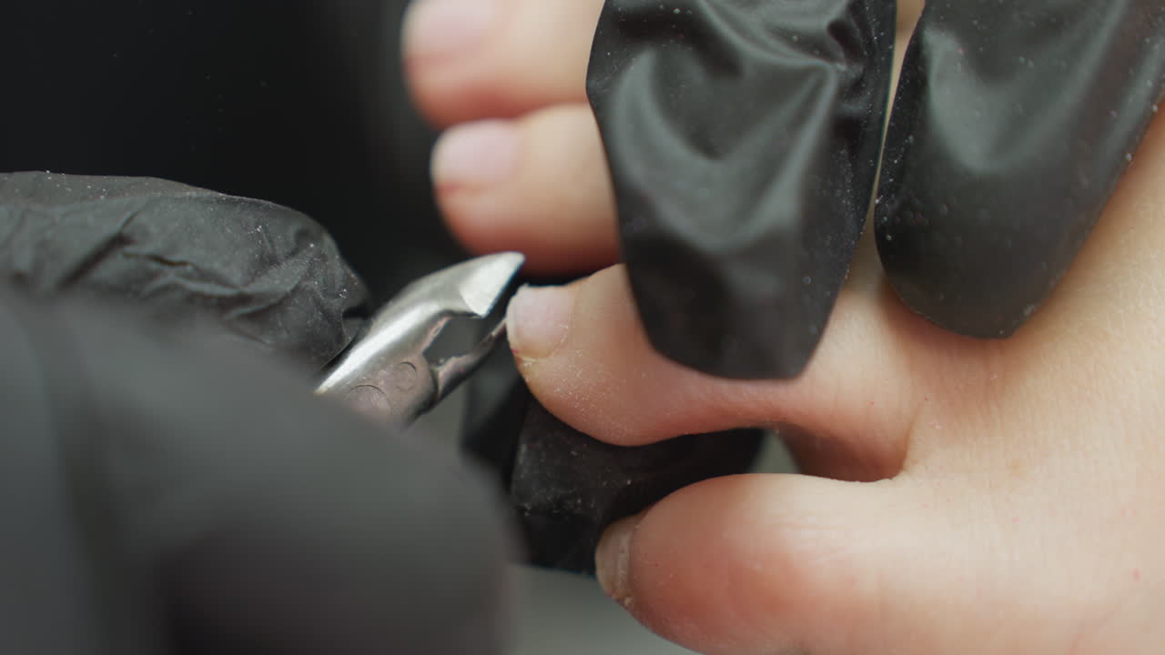 Close-up view of nail technician wearing black gloves using clipper to carefully trim client toenail during professional pedicure session, emphasizing precision, hygiene, and grooming process