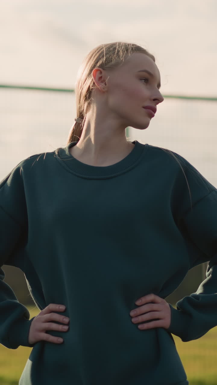 Giovane donna con maglione verde, mano sul fianco, si allena ruotando la testa da sinistra a destra, la luce del sole crea un bagliore dorato intorno a lei, con una vista parziale di qualcuno al suo fianco in un ambiente esterno