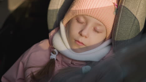 a young girl peacefully sleeping in a car seat, wearing a pink knit hat and scarf. the camera zooms in closely