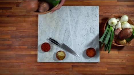vista desde arriba de las manos de un chef que prepara una mesa de madera y mármol con elementos para cocinar, verduras, condimentos, frijoles, cuchillo