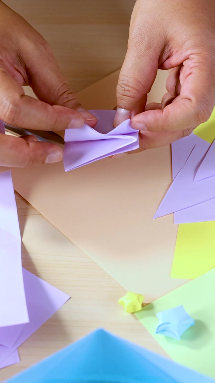 Una persona piega carta pastello per fare origami su un tavolo di legno, vista dall'alto, con illuminazione naturale brillante.
