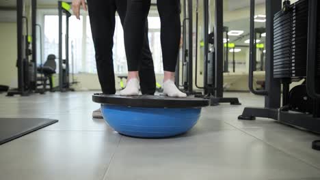a woman is balancing on an electric blue balance ball on the hardwood flooring of the gym, engaging her thigh and knee muscles for stability and building strength