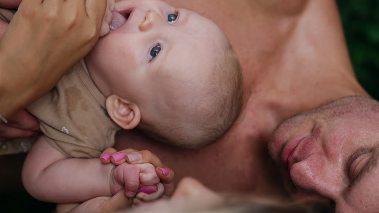 Male with naked torso holding a tiny baby. Grey-eyed infant puts his hands into mouth and mom wants to pull the hand out. Close up.